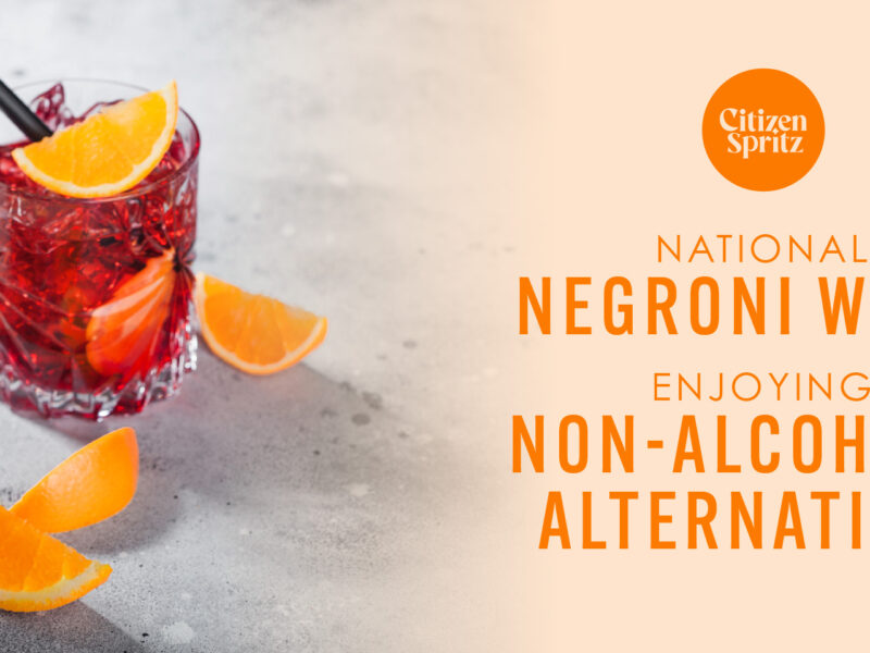 Two glasses of non-alcoholic Negroni garnished with orange slices and served with black straws, placed on a light grey surface. The text on the right side of the image reads 'Citizen Spritz - National Negroni Week: Enjoying Non-Alcoholic Alternatives' in orange and black letters.